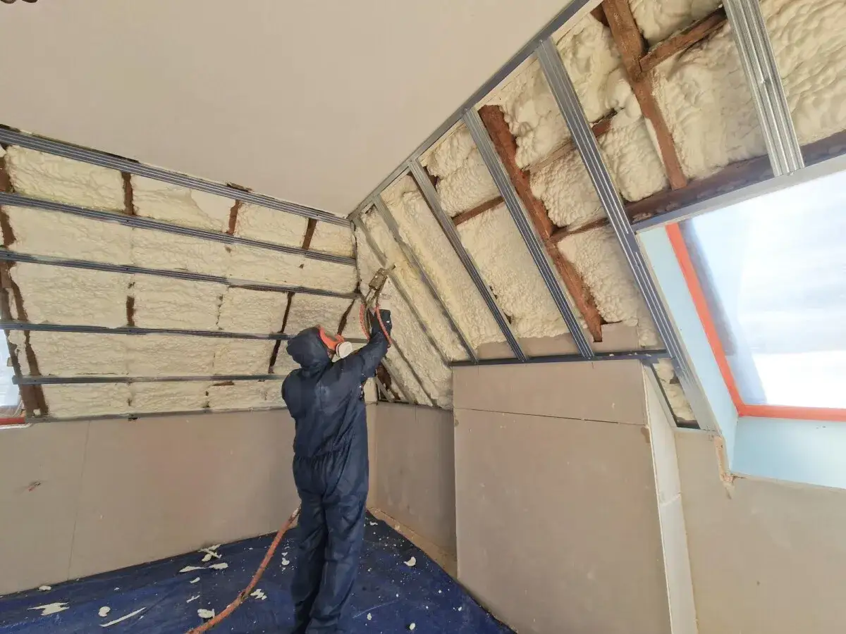 Un ouvrier en combinaison applique du polyur&eacute;thane projet&eacute; sur les murs et le toit d'une pi&egrave;ce en r&eacute;novation.