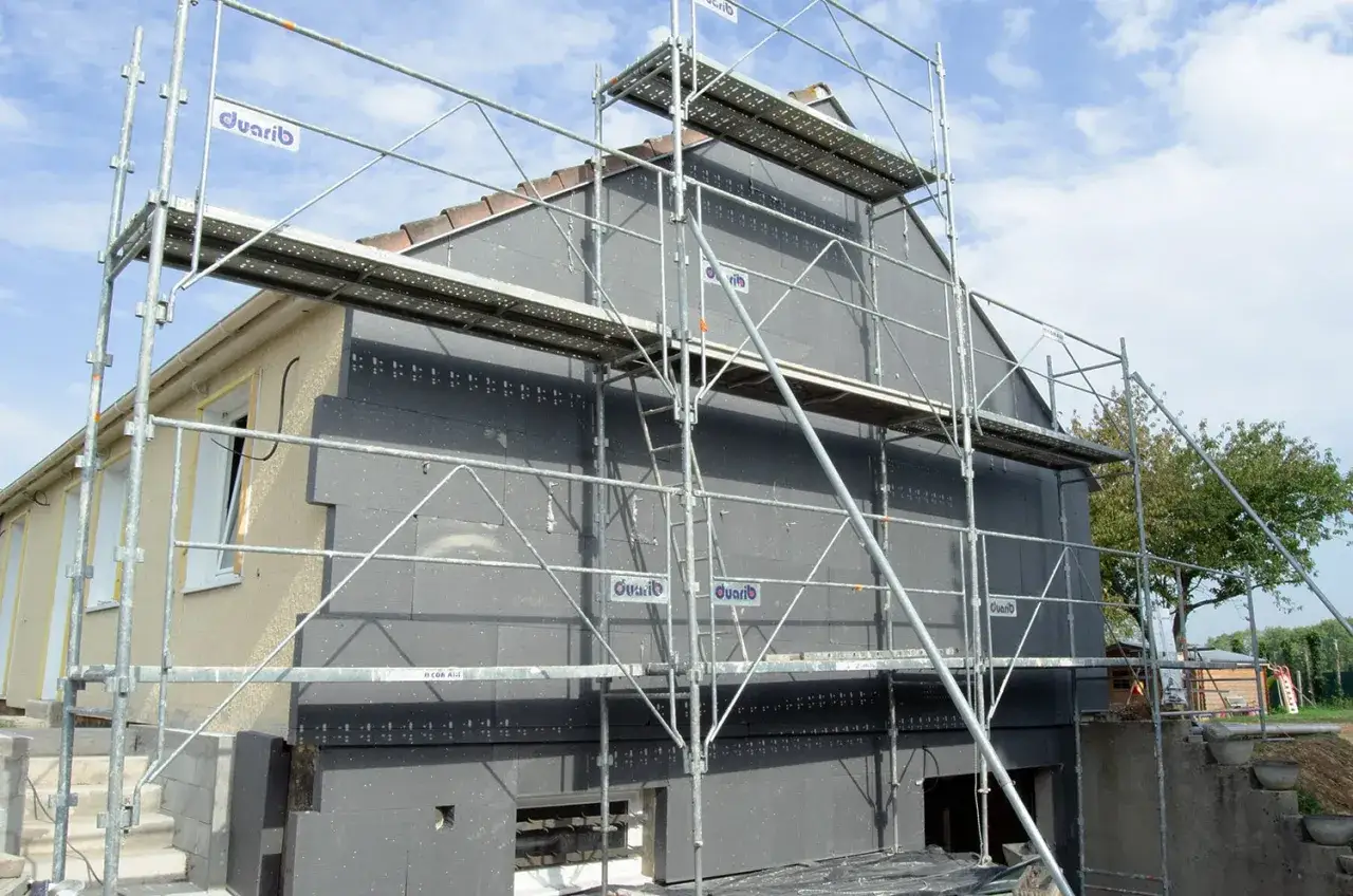 Chantier de ravalement avec isolation extérieure en cours sur une maison. Échafaudages métalliques entourant le bâtiment.