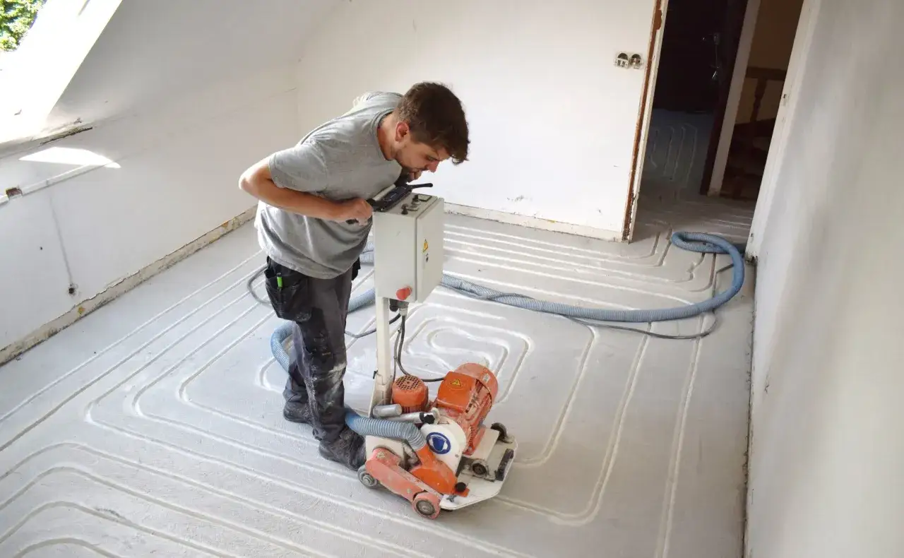 Un ouvrier utilise une ponceuse pour préparer un plancher rafraîchissant. Les tubes du système sont visibles sous la chape.