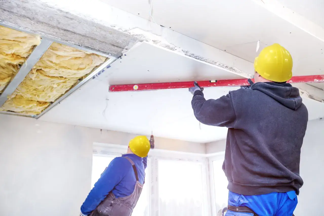 Pose de l'isolation du plafond d'un appartement. Des ouvriers en casque jaune installent des panneaux de plâtre et vérifient le niveau.