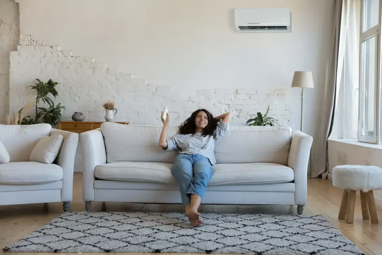 Femme souriante sur un canap&eacute;, t&eacute;l&eacute;commande &agrave; la main, profitant de la fra&icirc;cheur de la clim. Elle sait comment r&eacute;gler la clim en &eacute;t&eacute; pour un confort optimal.
