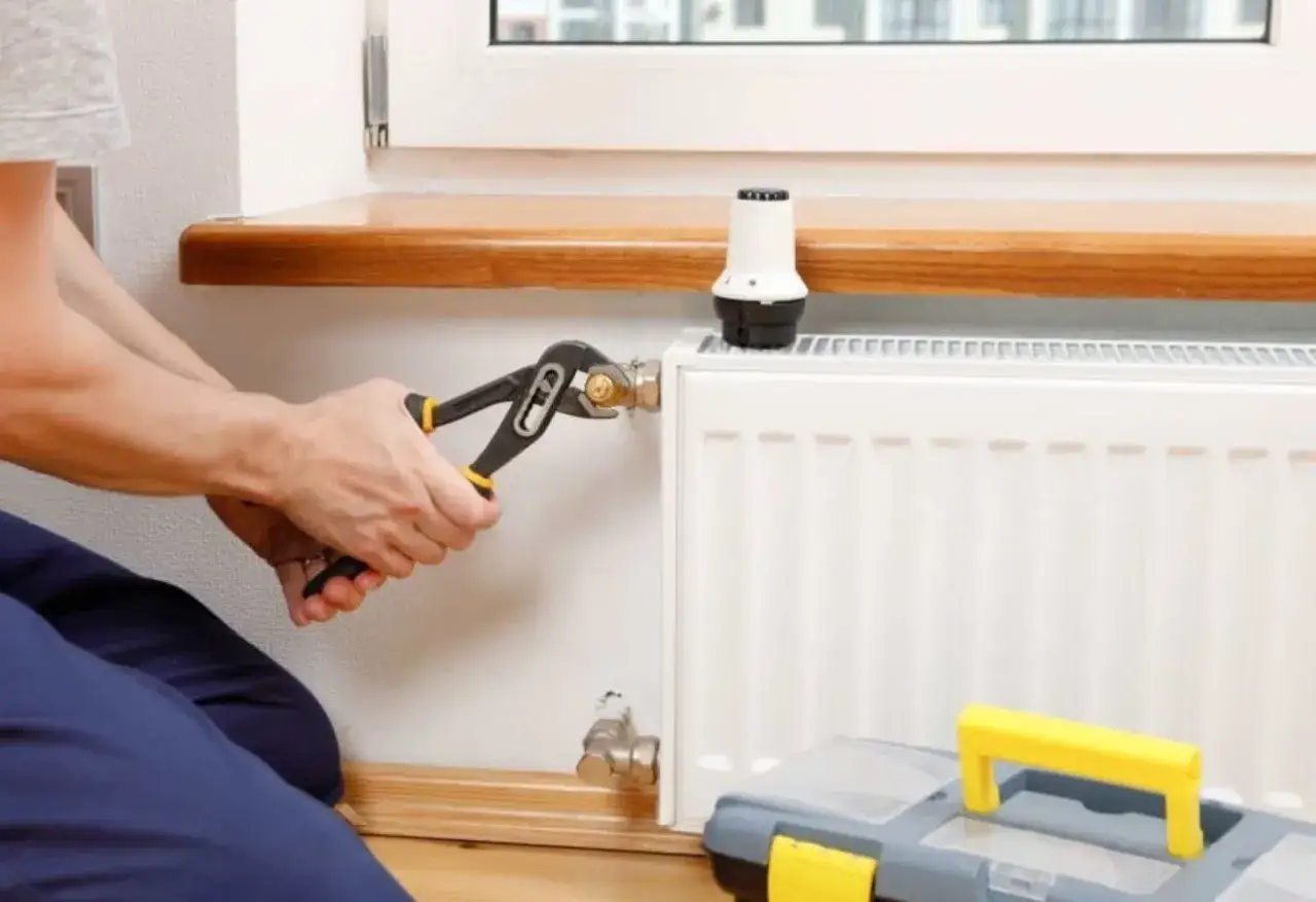 Un plombier utilise une pince pour effectuer le d&eacute;sembouage d'un radiateur. Une bo&icirc;te &agrave; outils est visible &agrave; proximit&eacute;.