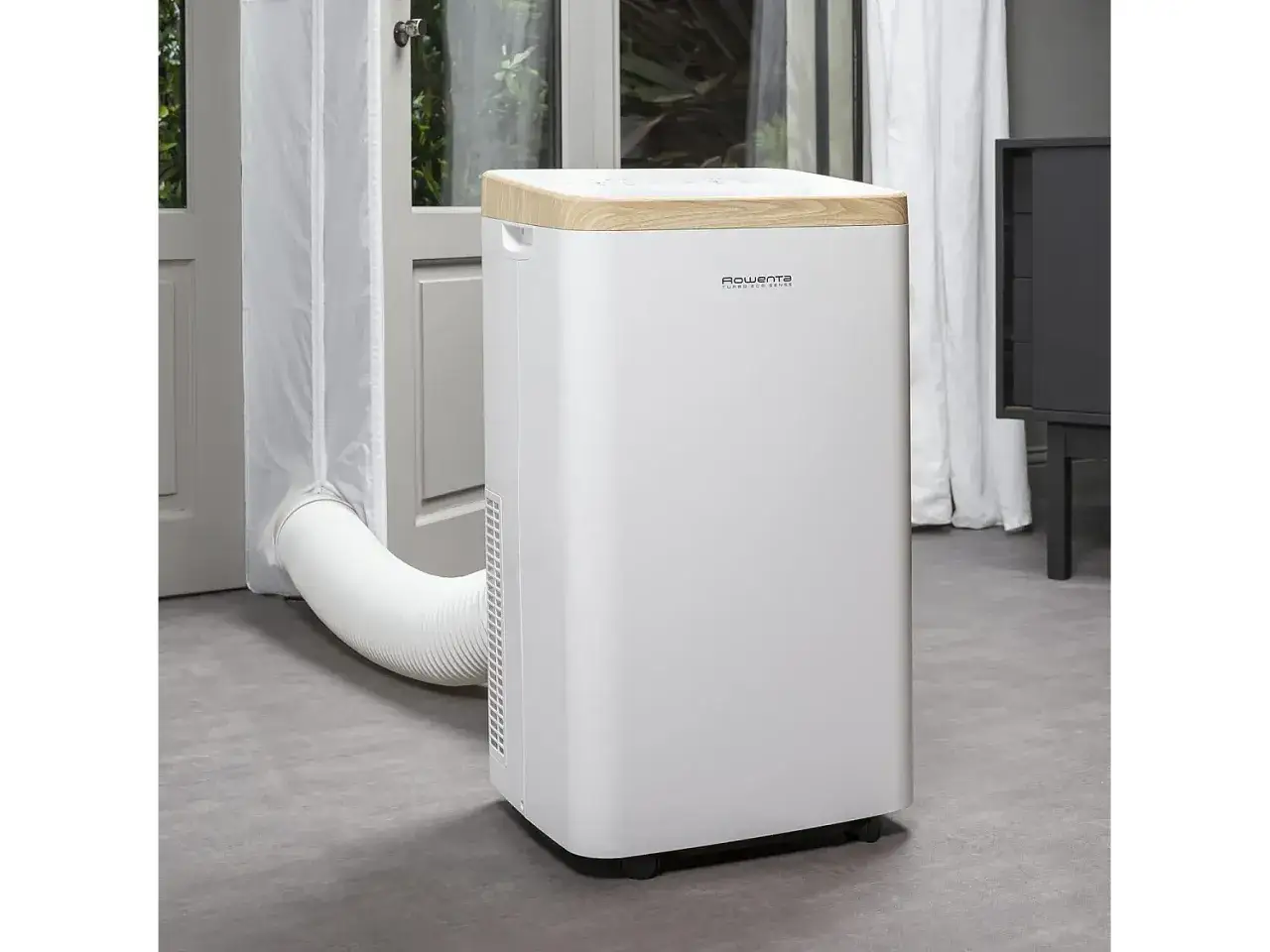 Climatiseur Rowenta blanc avec tuyau d'&eacute;vacuation passant par une porte ouverte. Design moderne avec finition bois&eacute;e.