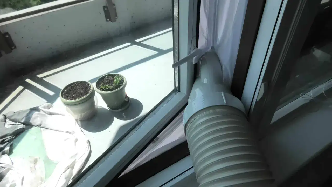 Tuyau d'un climatiseur mobile 80m2 sortant par une fen&ecirc;tre, avec des pots de plantes sur le rebord.