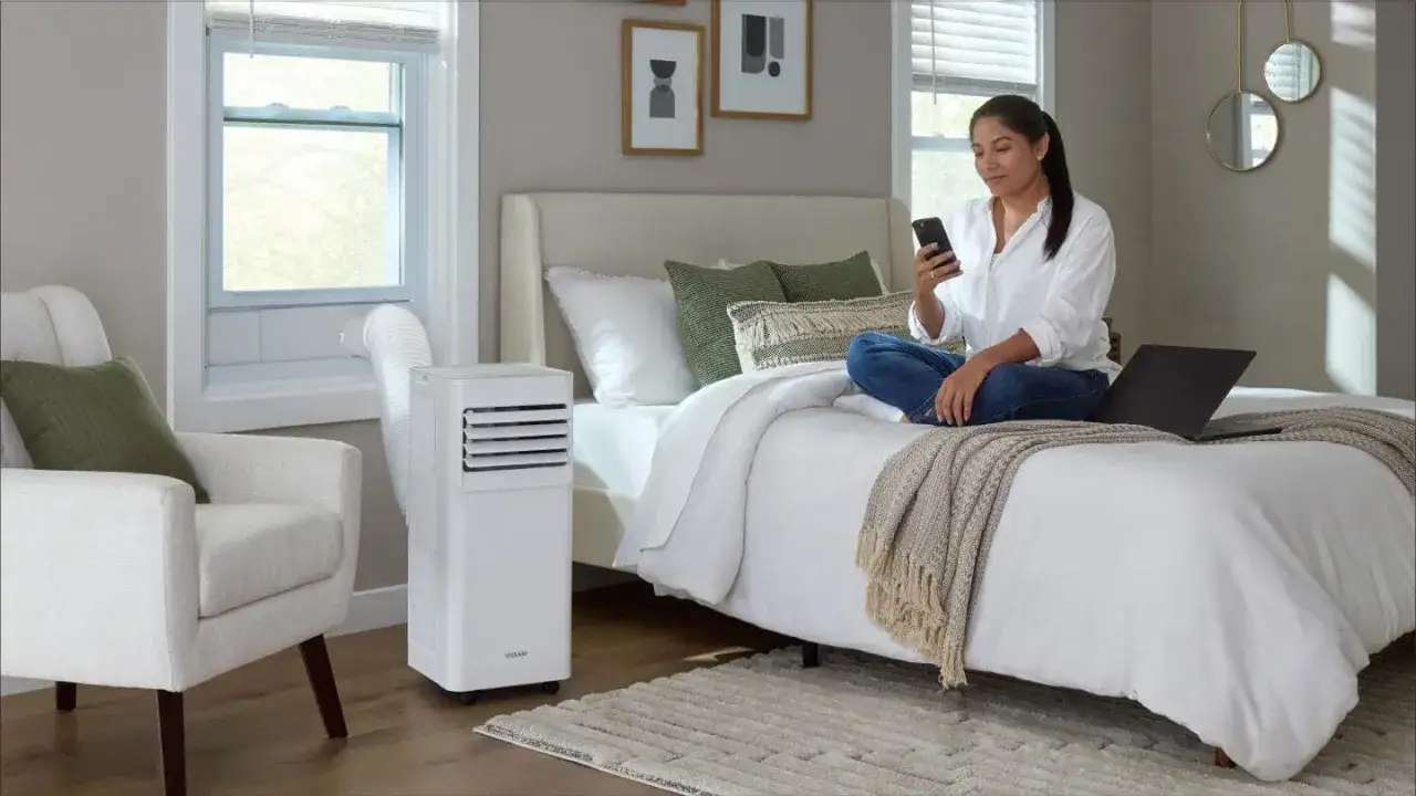Une femme utilise son t&eacute;l&eacute;phone sur un lit, &agrave; c&ocirc;t&eacute; d'un climatiseur mobile, la solution id&eacute;ale pour un confort optimal.