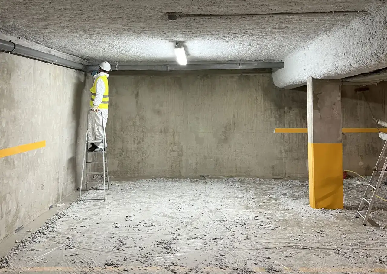 Un ouvrier en combinaison blanche et gilet jaune travaille sur un escabeau, appliquant du flocage isolation sur le plafond d'un parking.