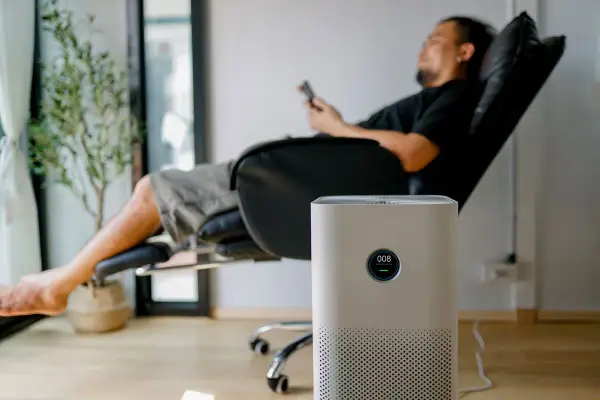 Un homme se repose dans un fauteuil, une plante verte à côté. Un purificateur d'air est au premier plan. Peut-on coucher une clim mobile ? Non, cet appareil est conçu pour rester debout.