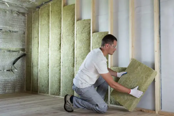 Un ouvrier installe des panneaux d'isolant minéral pour le **cout isolation interieur**. Le mur est en cours de construction, avec des montants en bois et une isolation déjà en place.