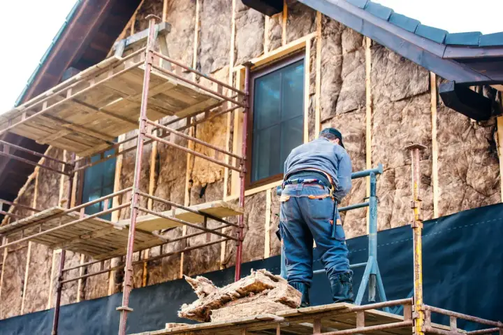 Un ouvrier sur un échafaudage installe l'isolation en fibre de bois sur le mur d'une maison en construction.