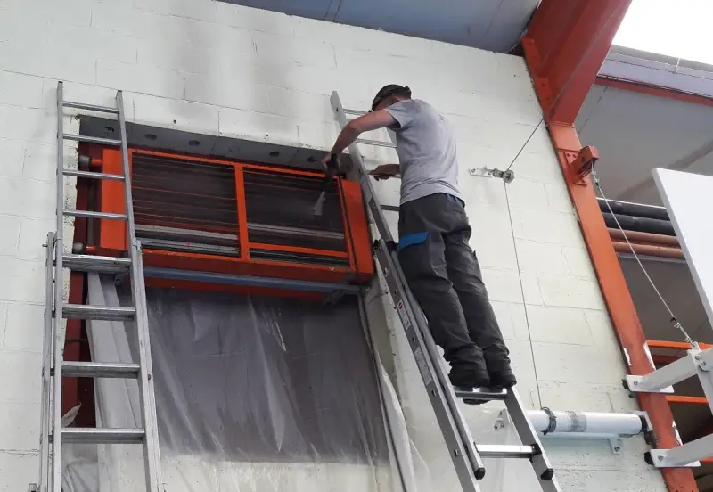 Un technicien sur une échelle nettoie un aérotherme de chauffage industriel.